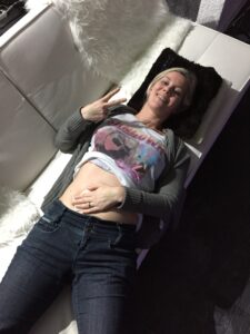 Christine Raab, eine junge Frau Anfang 30 mit blonden Haaren, liegt auf dem Rücken und zeigt mit den Händen das Victory Zeichen V. Ihr Shirt ist hochgeschoben und man sieht den Bauch. Sie hat eine Hand auf dem Bauch liegen. Der Bauch hat blaue Flecken von den vielen Spritzen