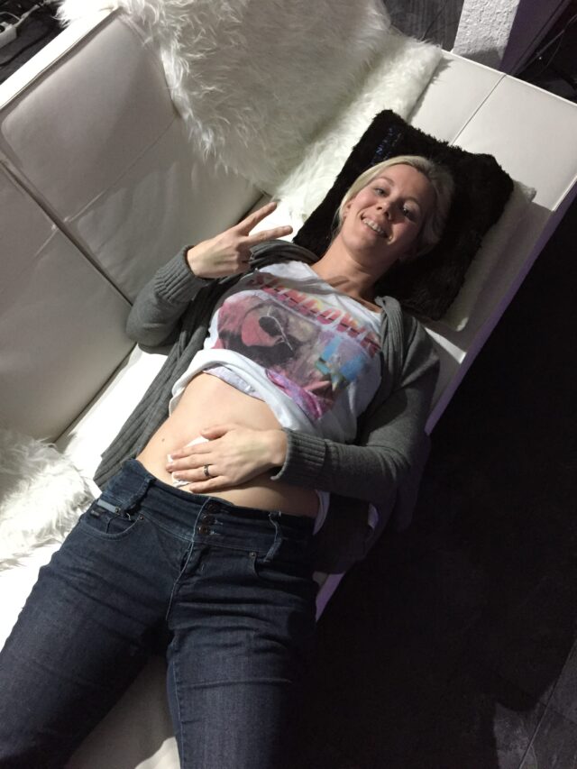 Christine Raab, eine junge Frau Anfang 30 mit blonden Haaren, liegt auf dem Rücken und zeigt mit den Händen das Victory Zeichen V. Ihr Shirt ist hochgeschoben und man sieht den Bauch. Sie hat eine Hand auf dem Bauch liegen. Der Bauch hat blaue Flecken von den vielen Spritzen