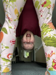 Christine, eine Frau Mitte 40, steht in einer leicht gekrätschten Position. Sie hat den Oberkörper nach vorne gebeugt und schaut zwischen den Beinen durch in die Kamera. Sie hat den Mund auf als würde sie schreien. Sie trägt eine Leggins mit grün und orangenem Muster und ein weinrotes Oberteil.